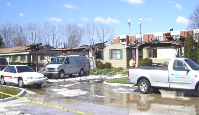 The aftermath of a house fire, with damaged buildings and emergency response vehicles present